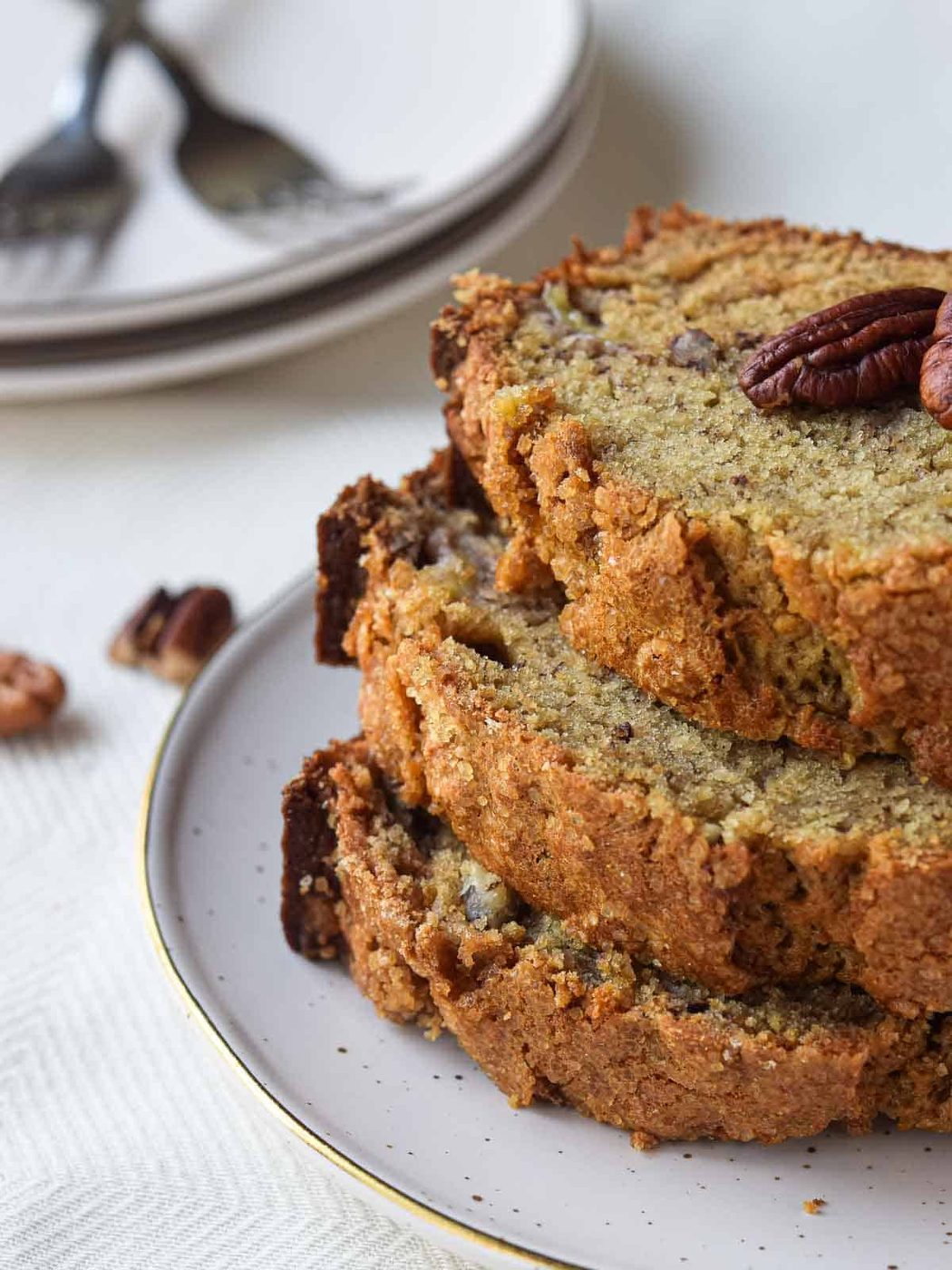 Three slices of banana bread stacked on a plate with pecans in the background.