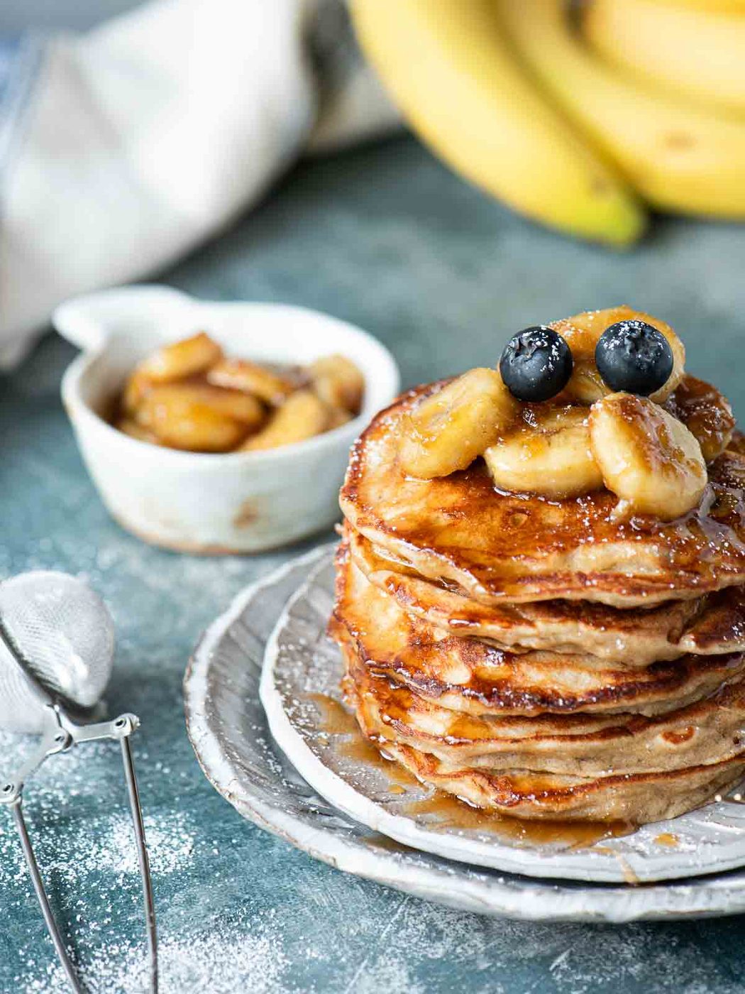 A stack of pancakes with bananas on top placed on grey plate over a blue surface.