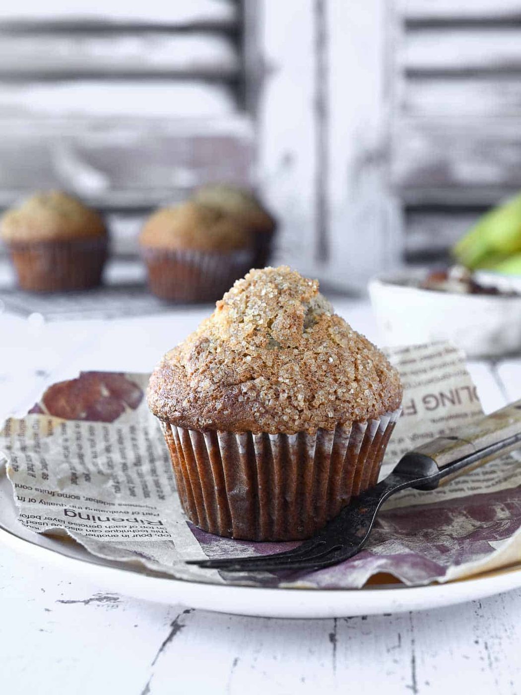 Banana muffin on a white plate with muffins and pecans in the background.