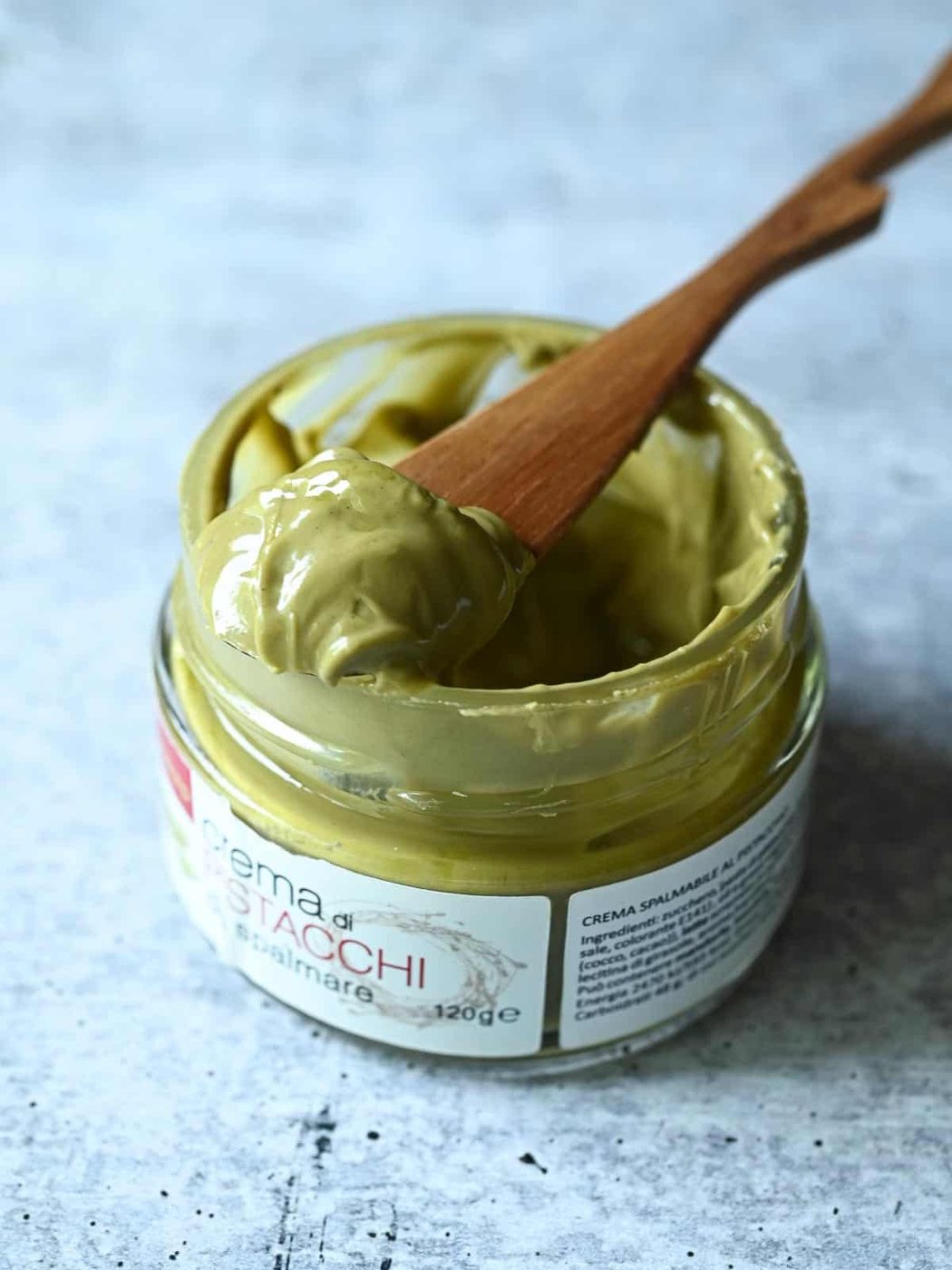 A spoon dipped into a jar of pistachio cream on a grey surface.