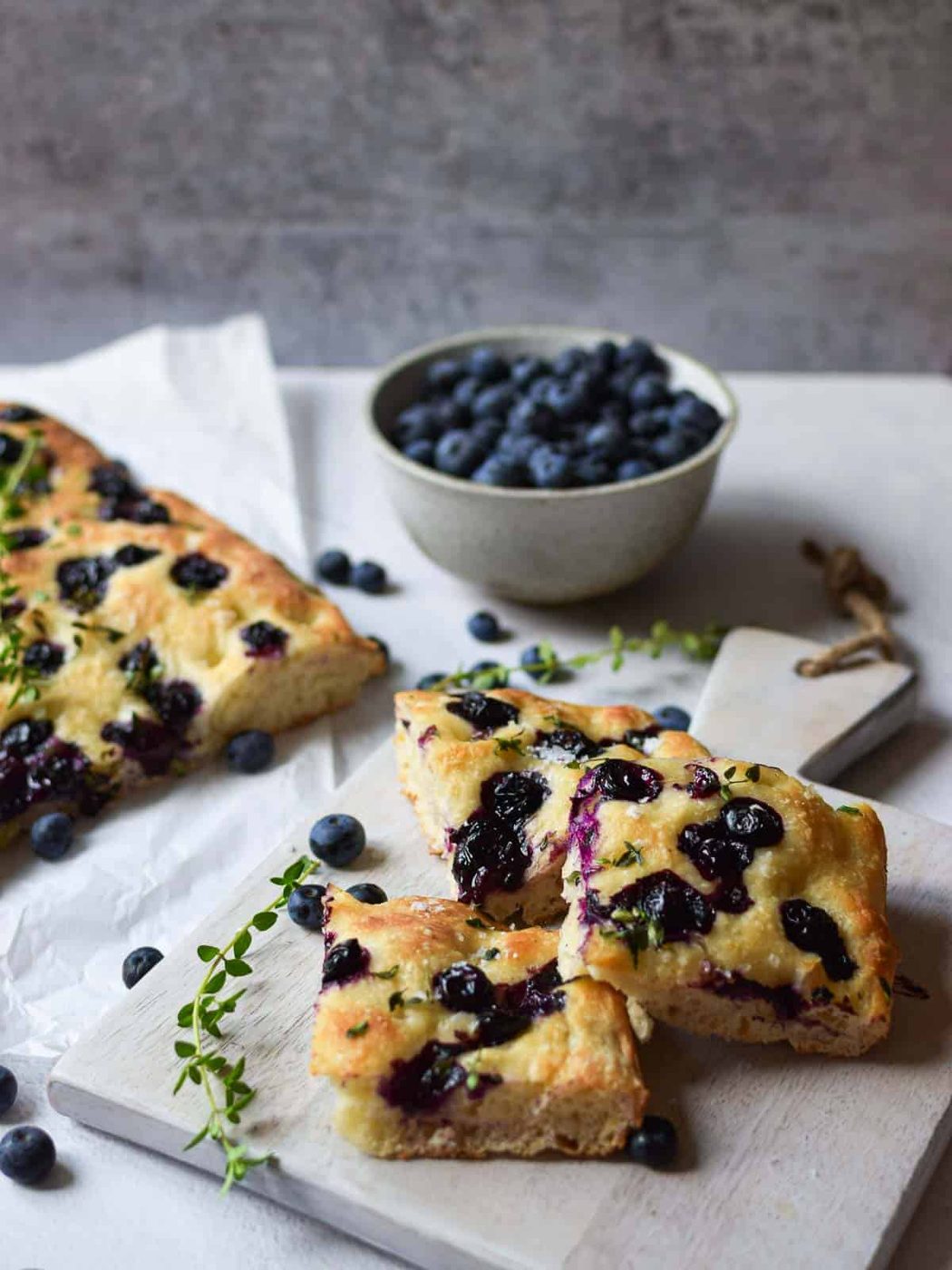 Angled view of blueberry thyme focaccia on a grey cutting board with blueberries in the background.