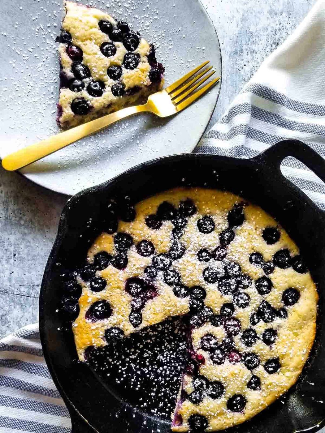 Overhead view of blueberry pancake skillet with a slice cut out on a plate in a black skillet