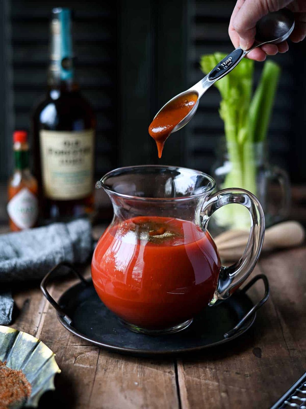 A hand adding barbeque sauce to a pitcher of bloody mary mix.