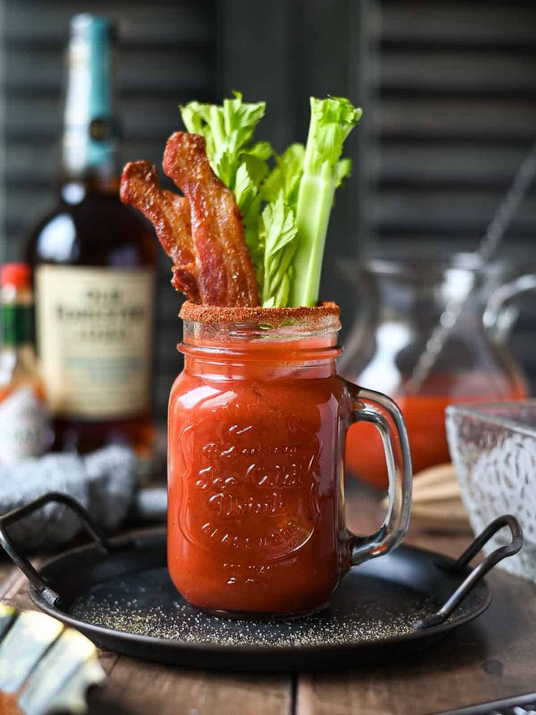 Eye-level view of a Bourbon Bloody Mary, plus Bacon and celery in a clear Mason jar.