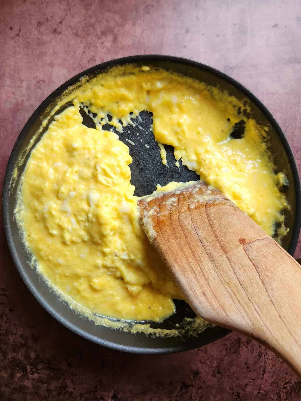 A wooden spoon shown moving scrambled eggs in a pan.