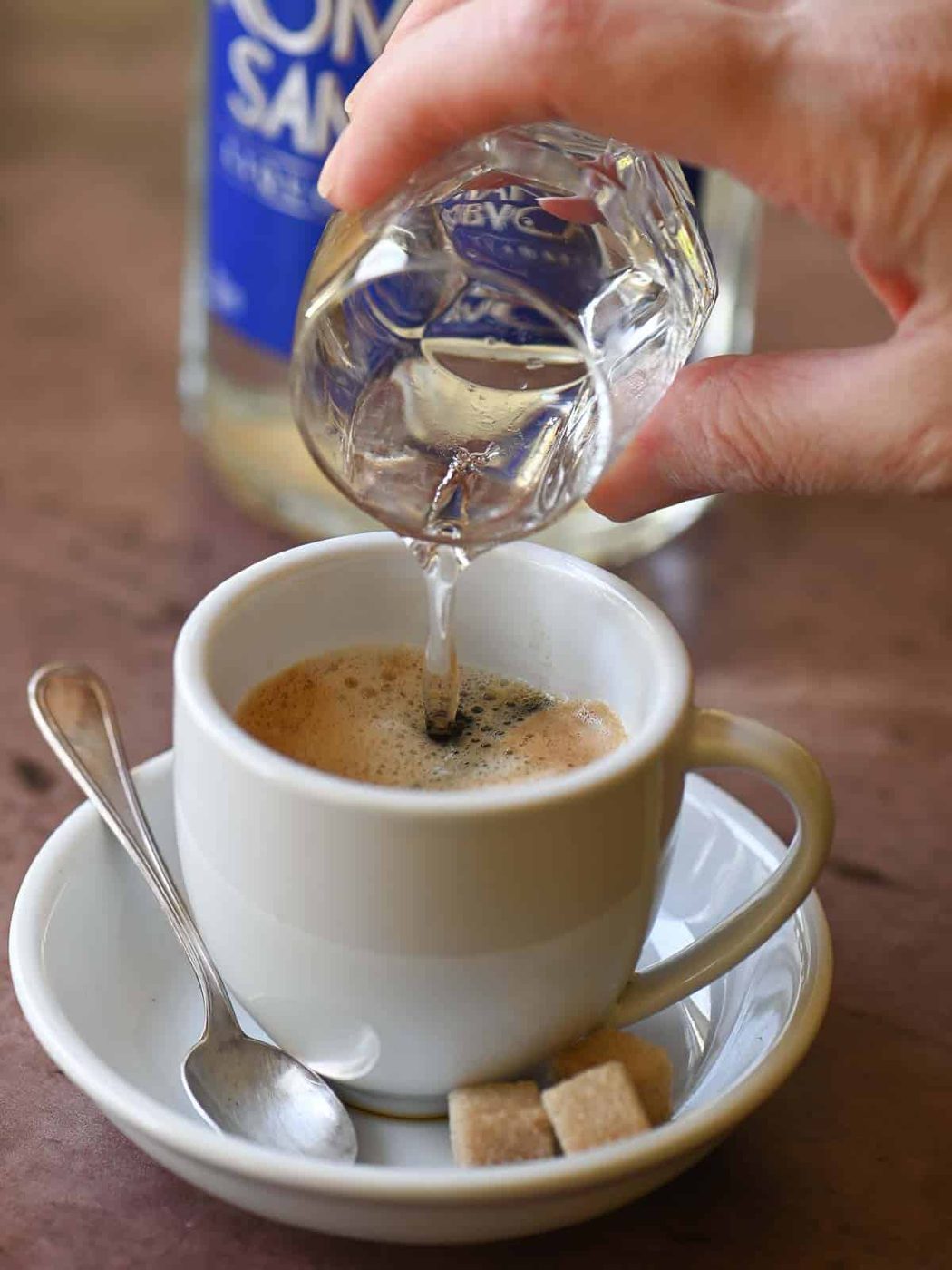 A hand pouring Sambuca into a white cup with espresso.