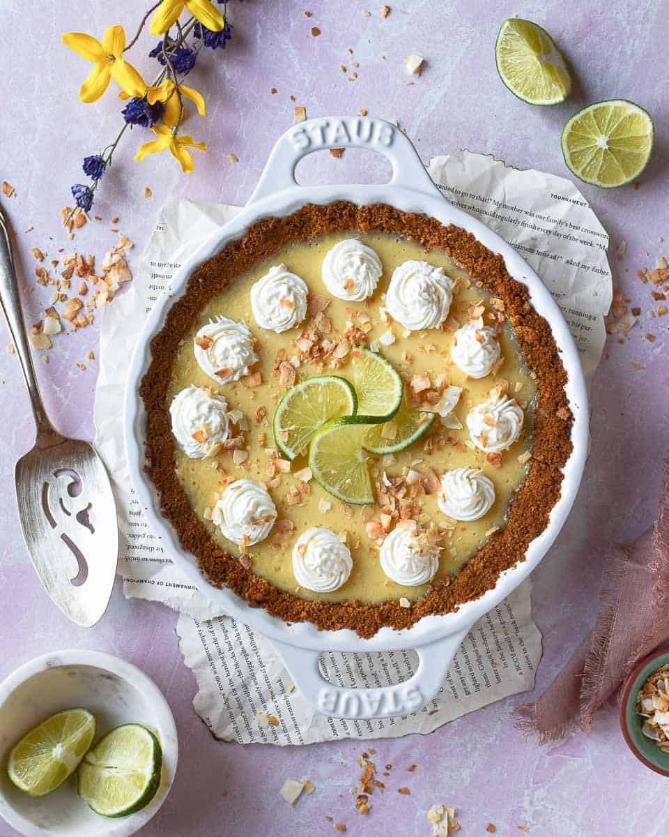 Overhead shot of coconut key lime pie in white pie dish placed on a pink background. Scattered limes in eh background.