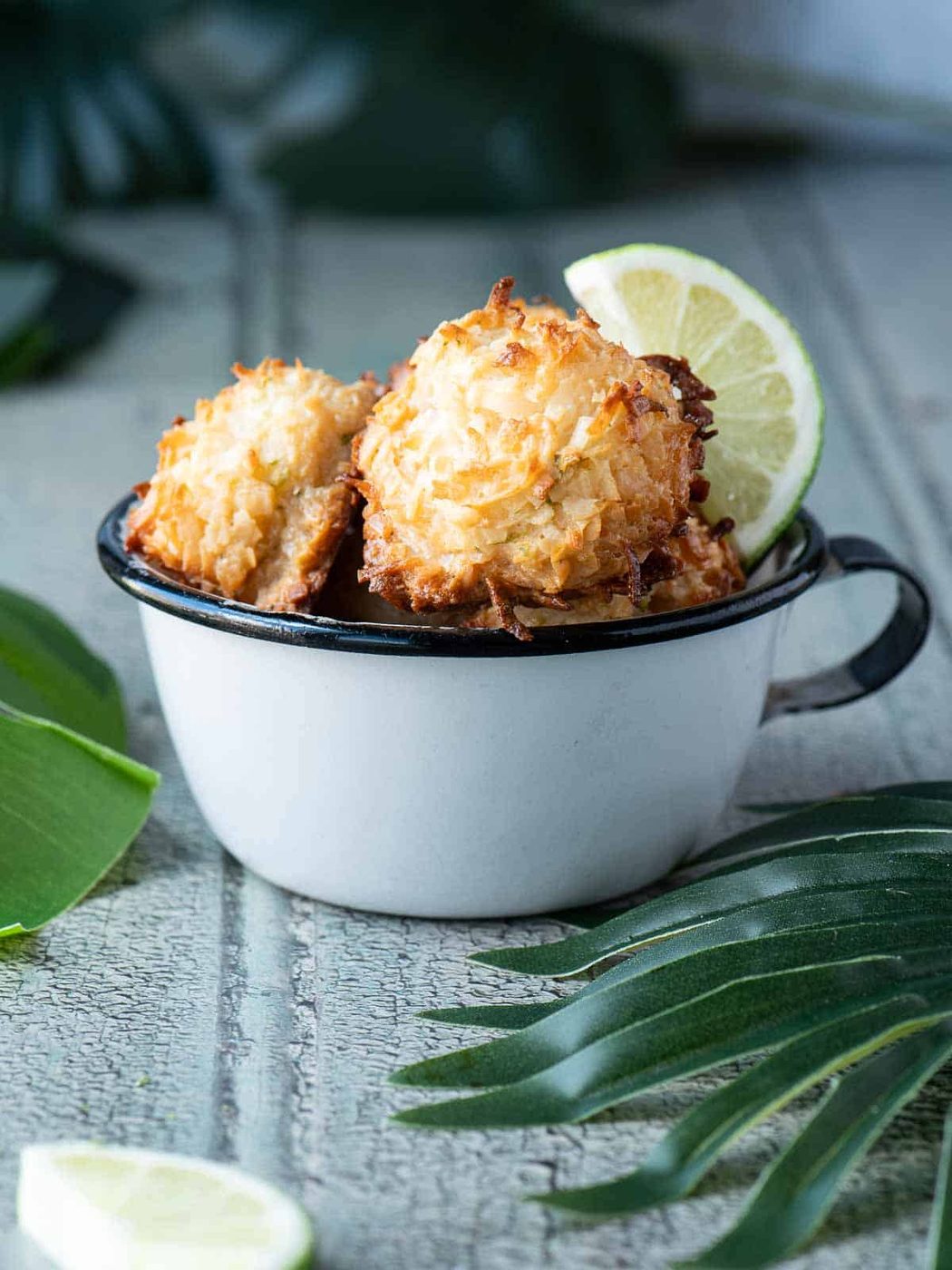 Coconut macaroons in a white cup with palm leaves and limes next to it.