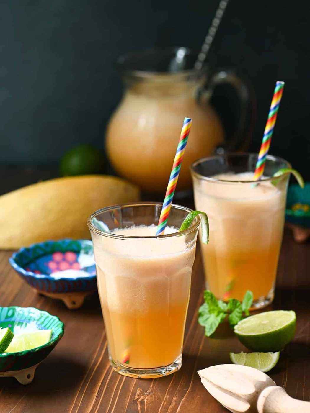 Two clear glasses of cantaloupe drink with straws on a wooden board with limes.