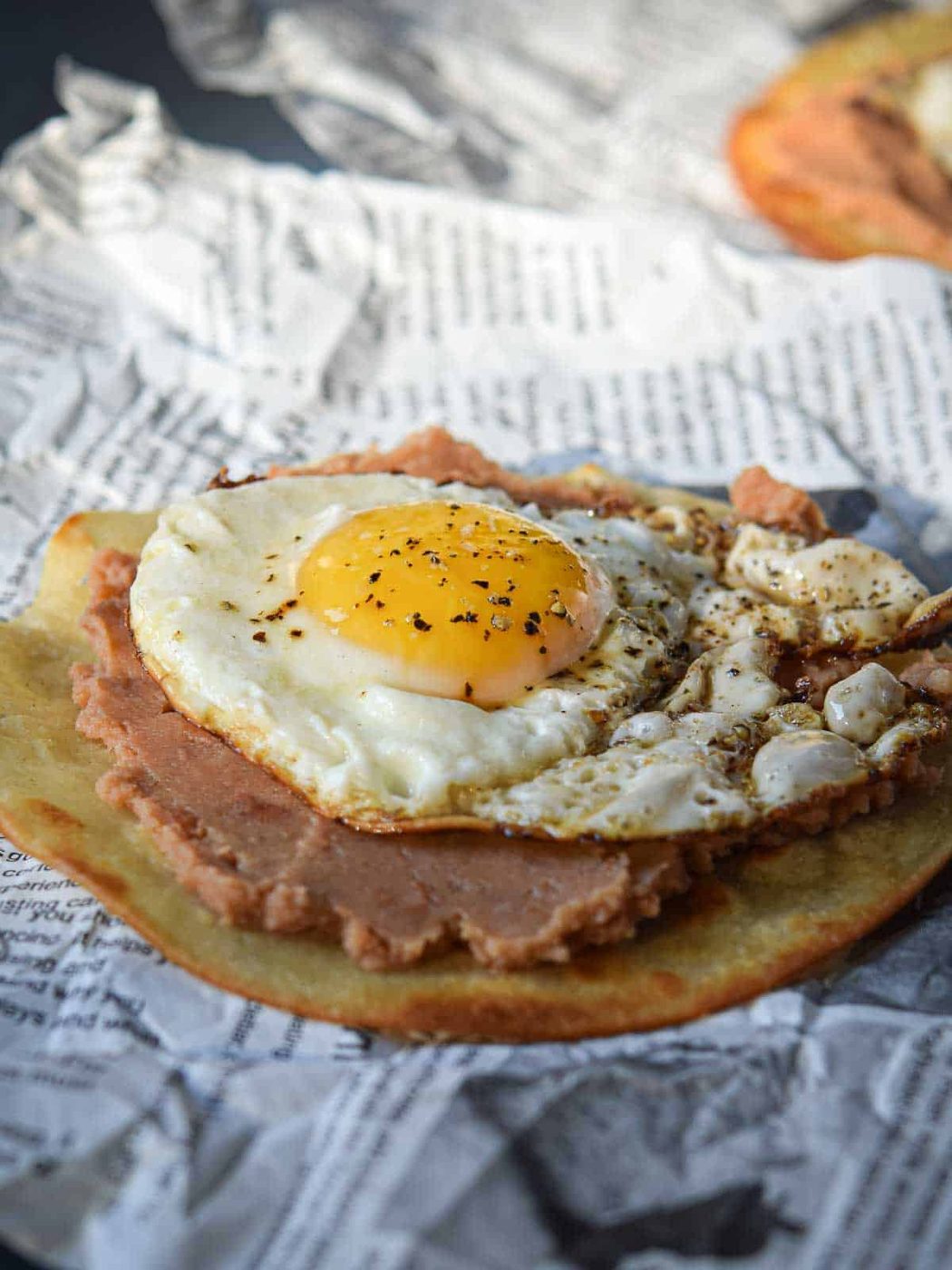 Fried egg on top of a fried tortilla with refried beans over newspaper.