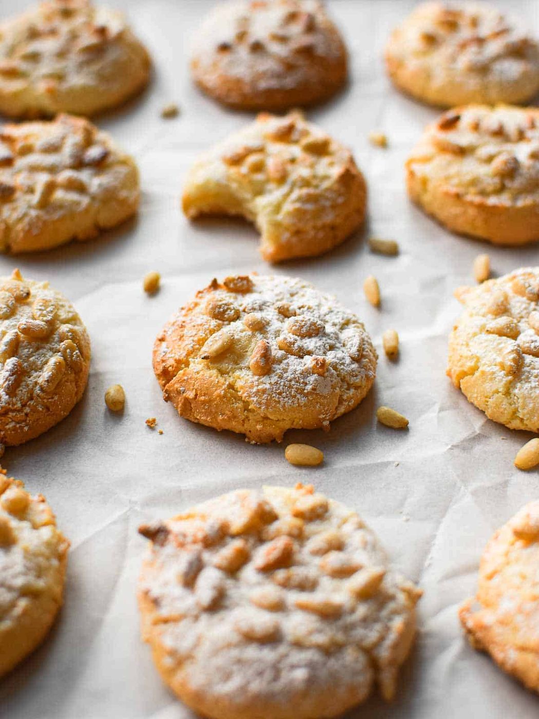 Angled view of baked Italian pignoli cookies on a baking sheet covered with white parchment paper. One cookie has a bite taken out.