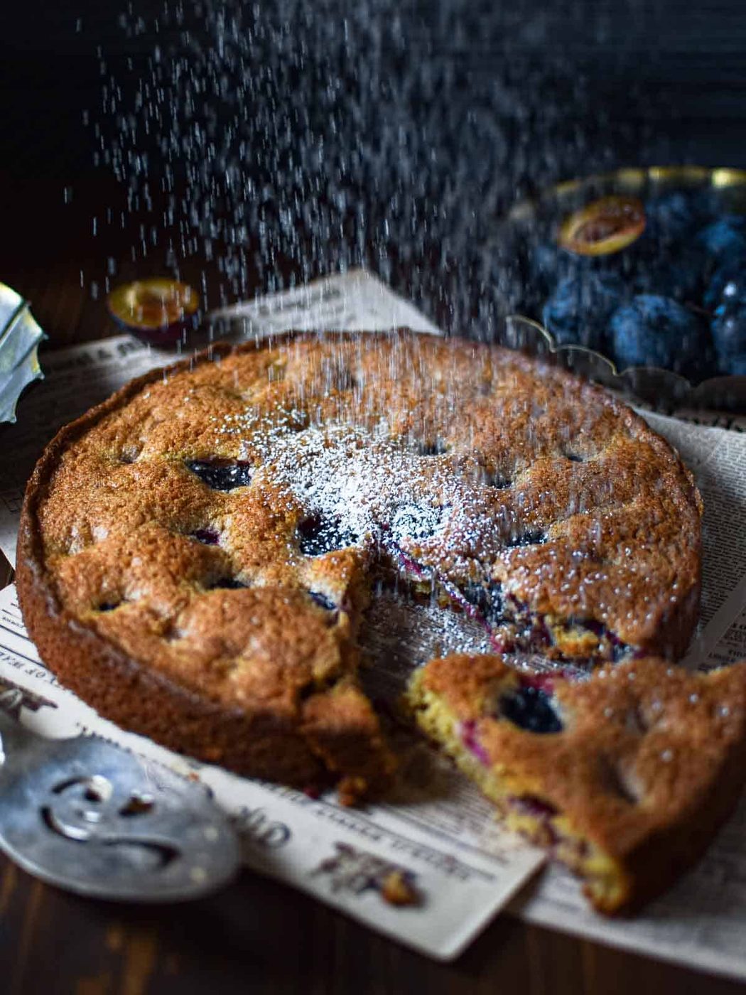 Angled view of plum cake with a slice removed while being dusted with powdered sugar on a wood surface.