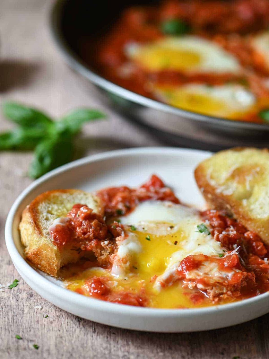 Savory eggs in tomato sauce with garlic bread on a white plate, fresh basil garnish.