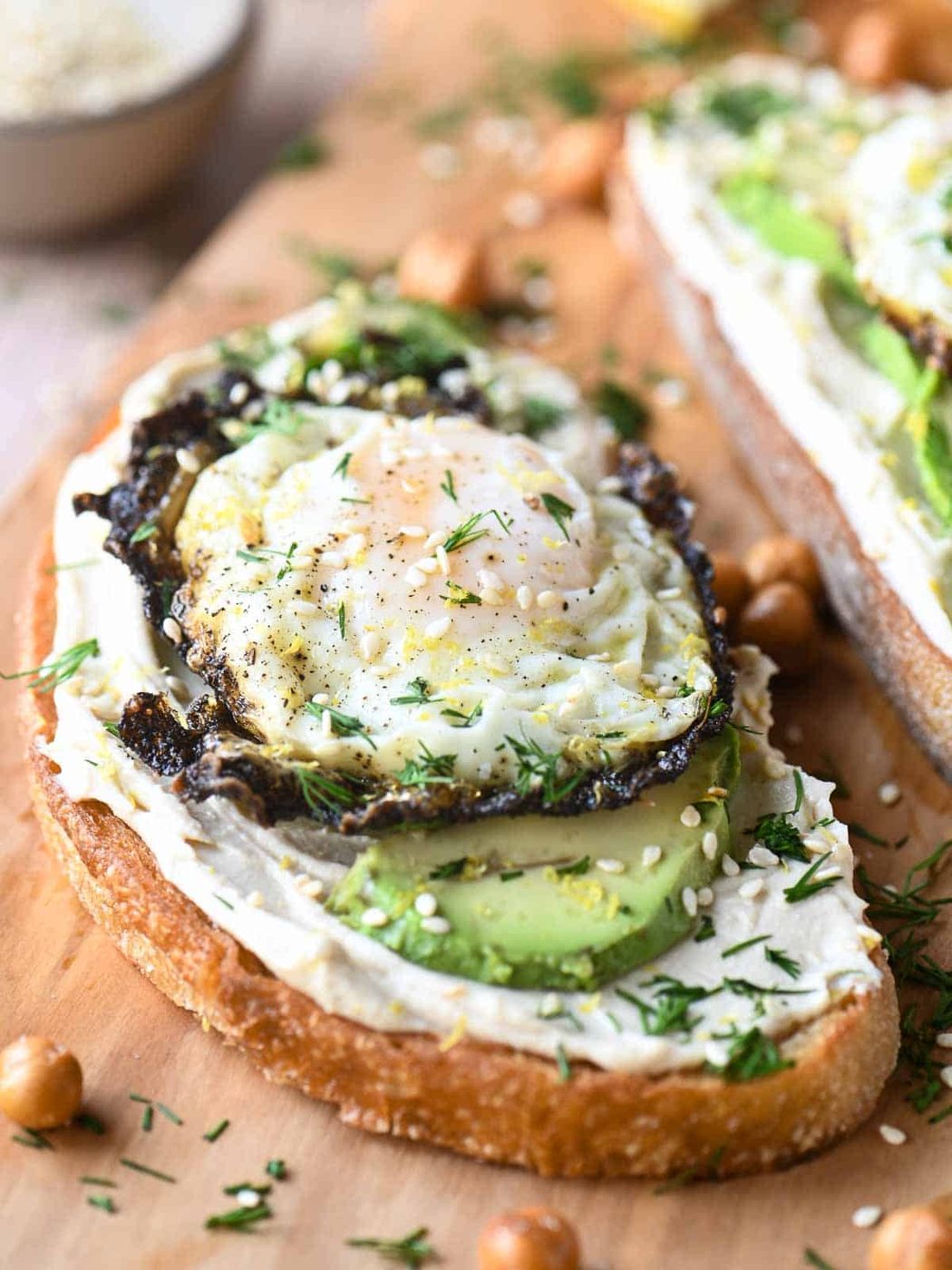 Hummus toast on a cutting board with eggs, avocado and dried chick peas.