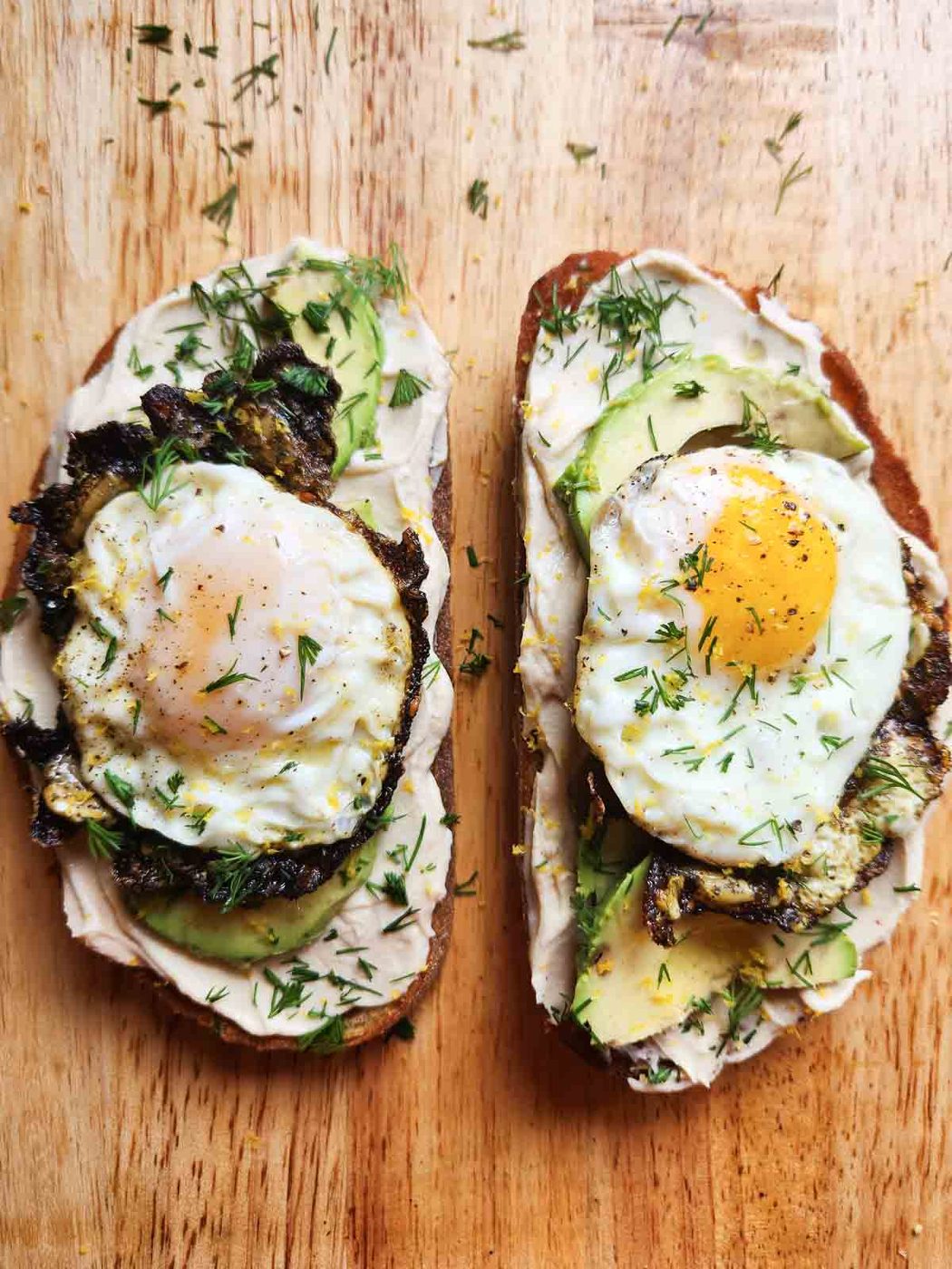 Two slices of hummus toast with fried eggs on a wooden board.