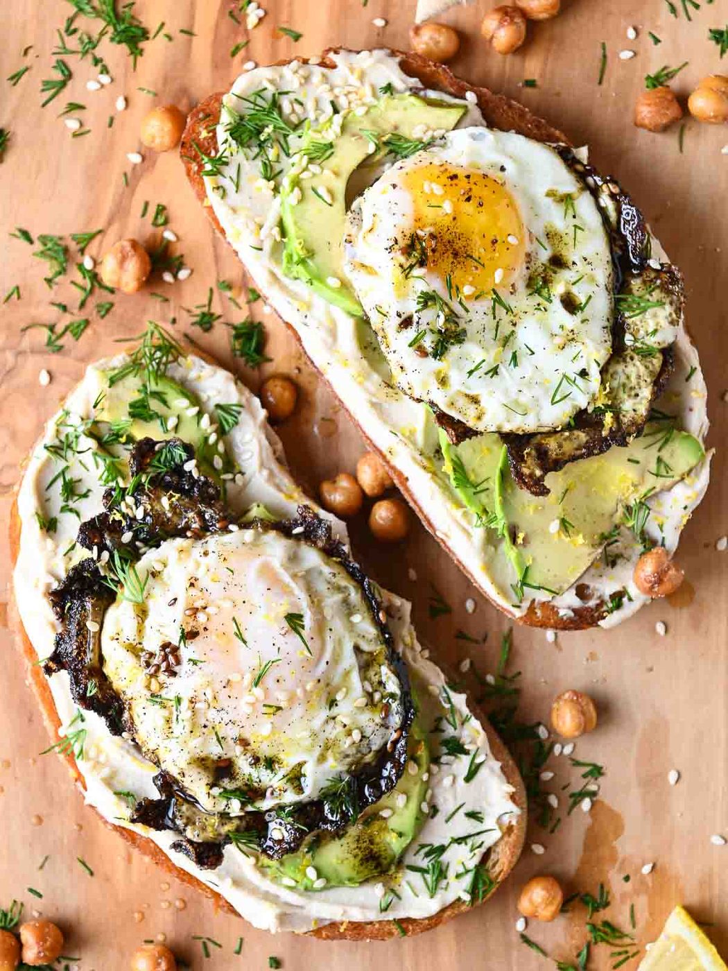 Overhead of hummus toasts on a wooden cutting board with fried eggs and avocado.