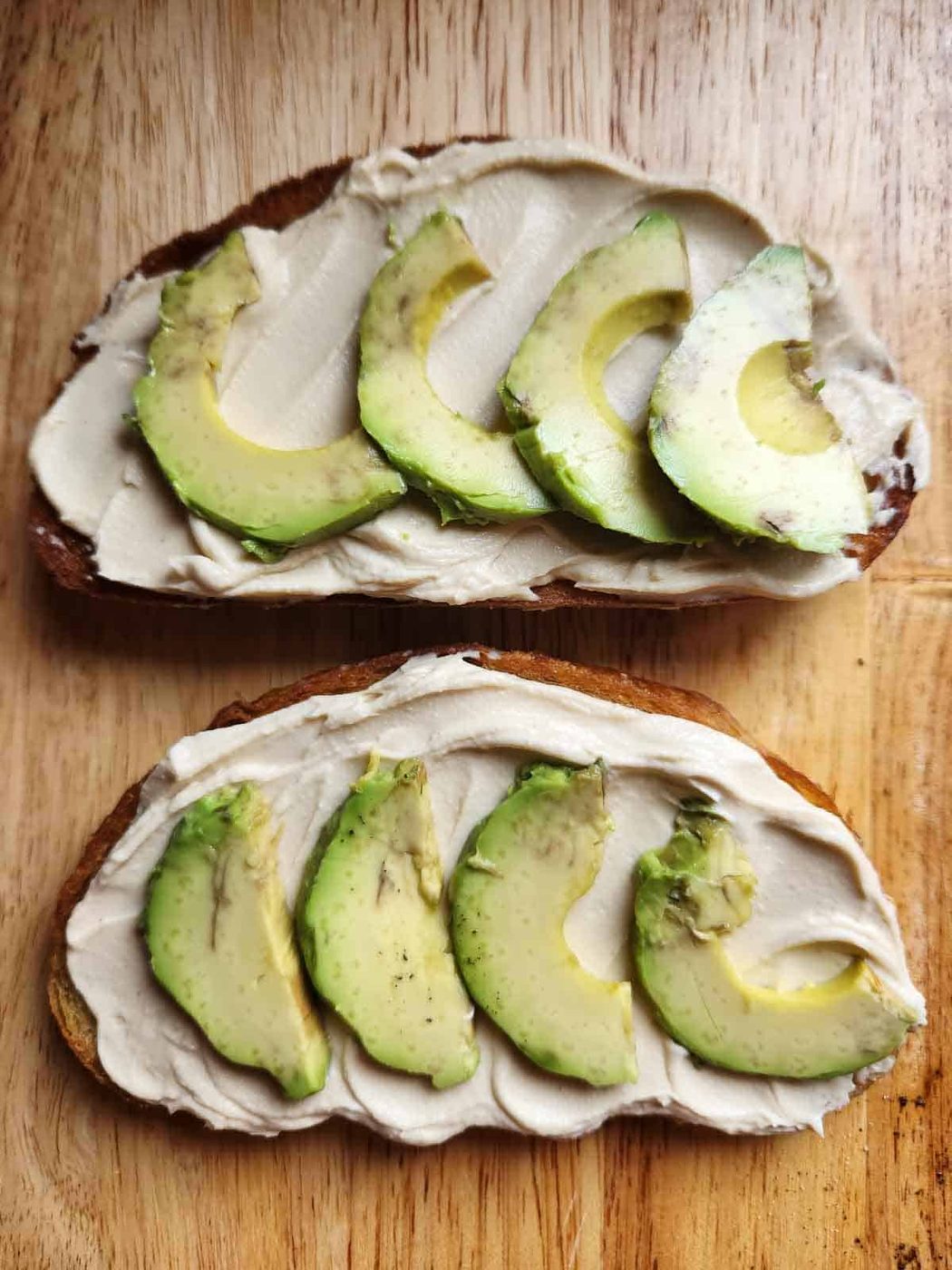 Two slices of bread with hummus and avocado on a wooden board.