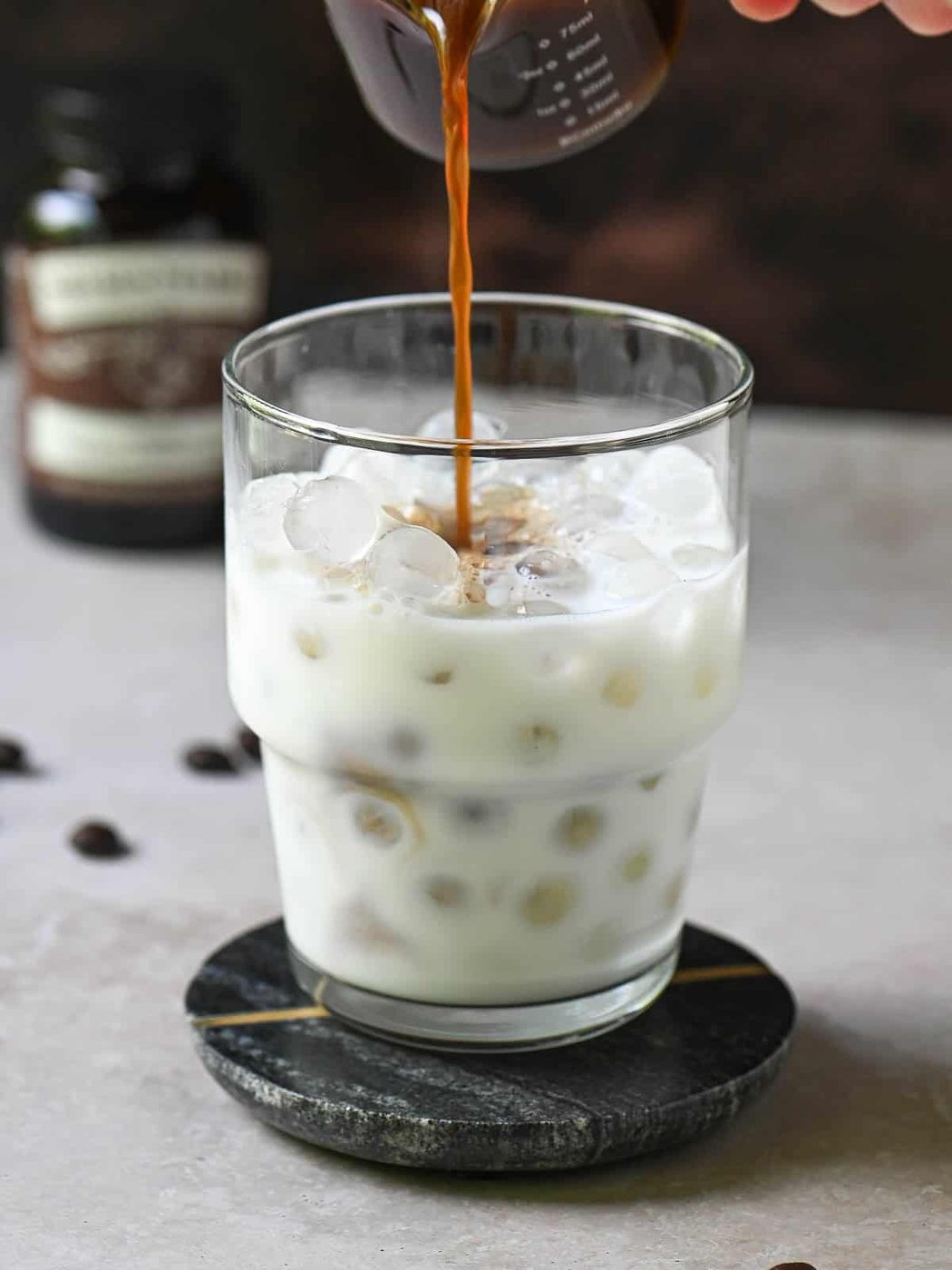 Coffee is being poured into a clear glass with milk and ice.