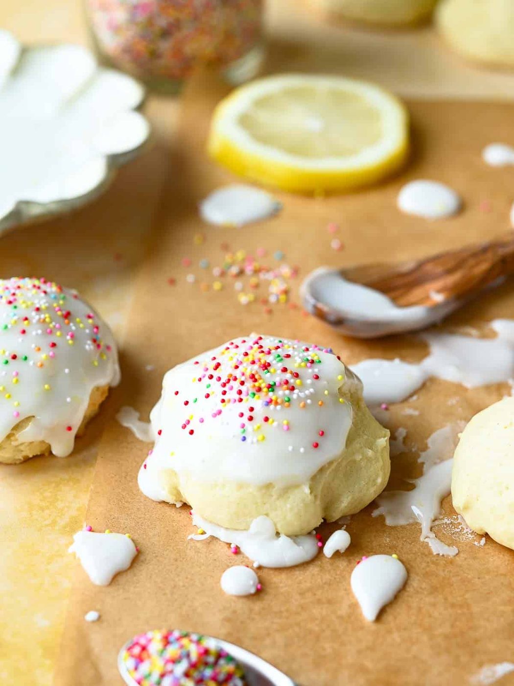 Italian lemon drop cookies with icing and sprinkles on parchment paper with lemons and sprinkles in the background.