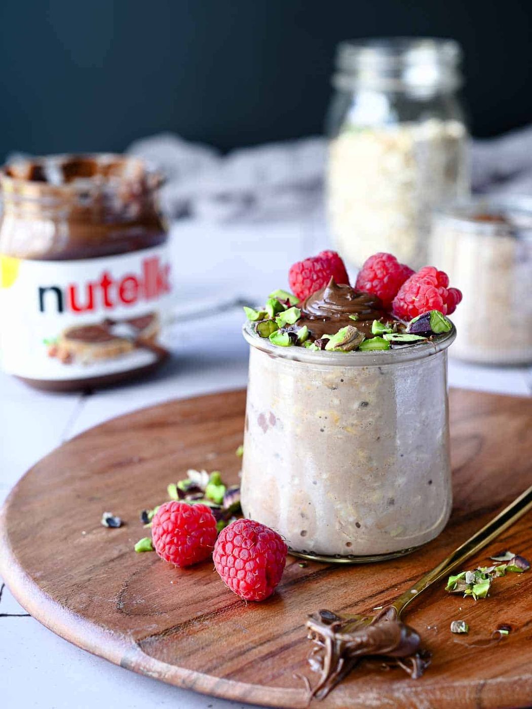 Nutella overnight oats in a clear jar with raspberries and pistachios on wood.