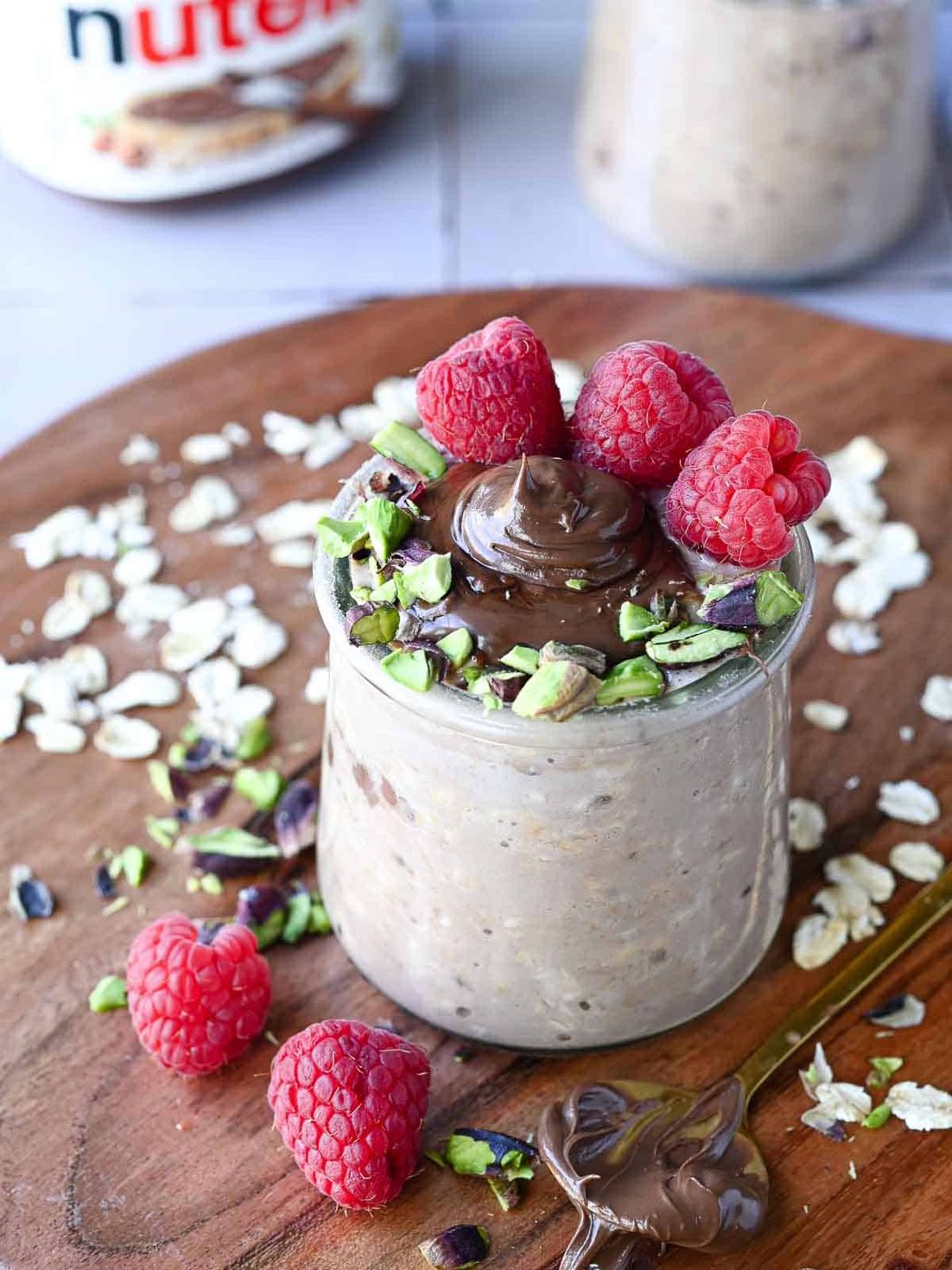 Nutella overnight oats in a clear jar with raspberries and pistachios on wood.