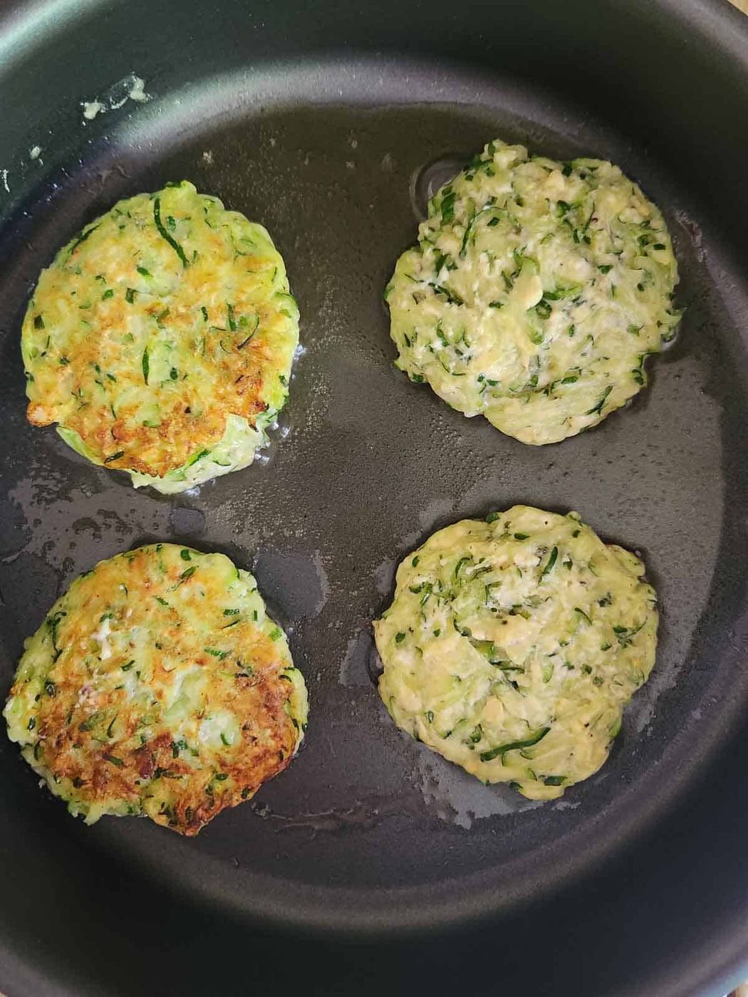 Four zucchini pancakes in a skillet being cooked.