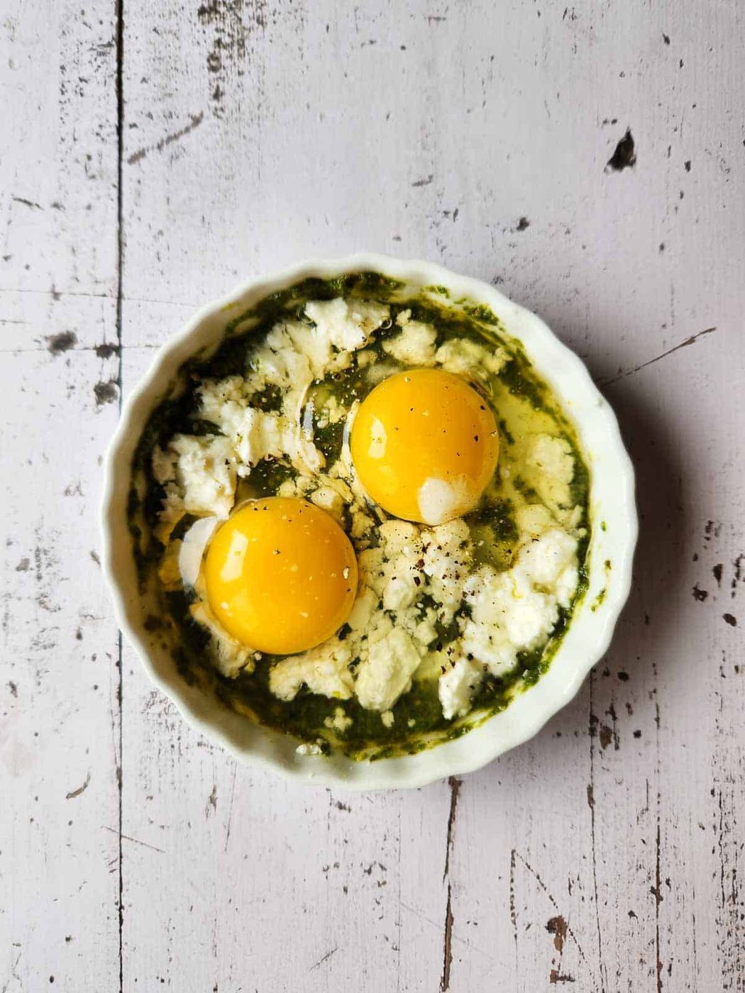 Pesto, feta, cream, and two eggs in a white ramekin.