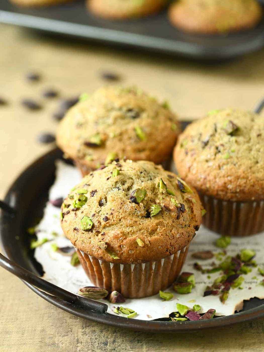 Three pistachio muffins on a black tray with scattered pistachios.