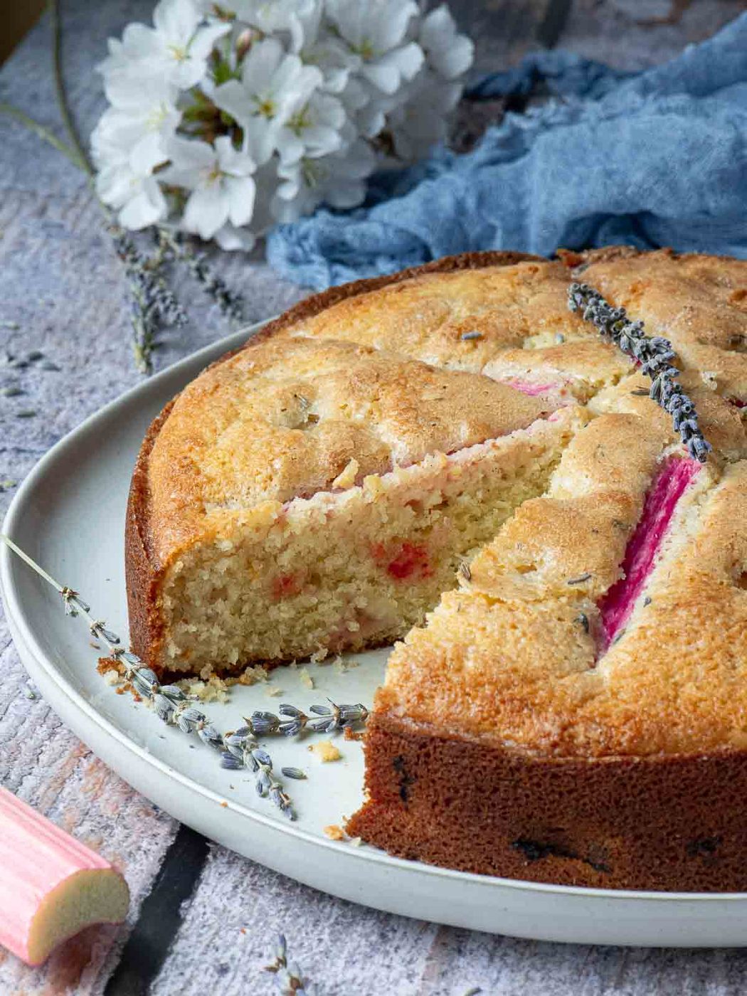 Rhubarb lavender cake with a slice removed on a wood surface with flowers in the back.