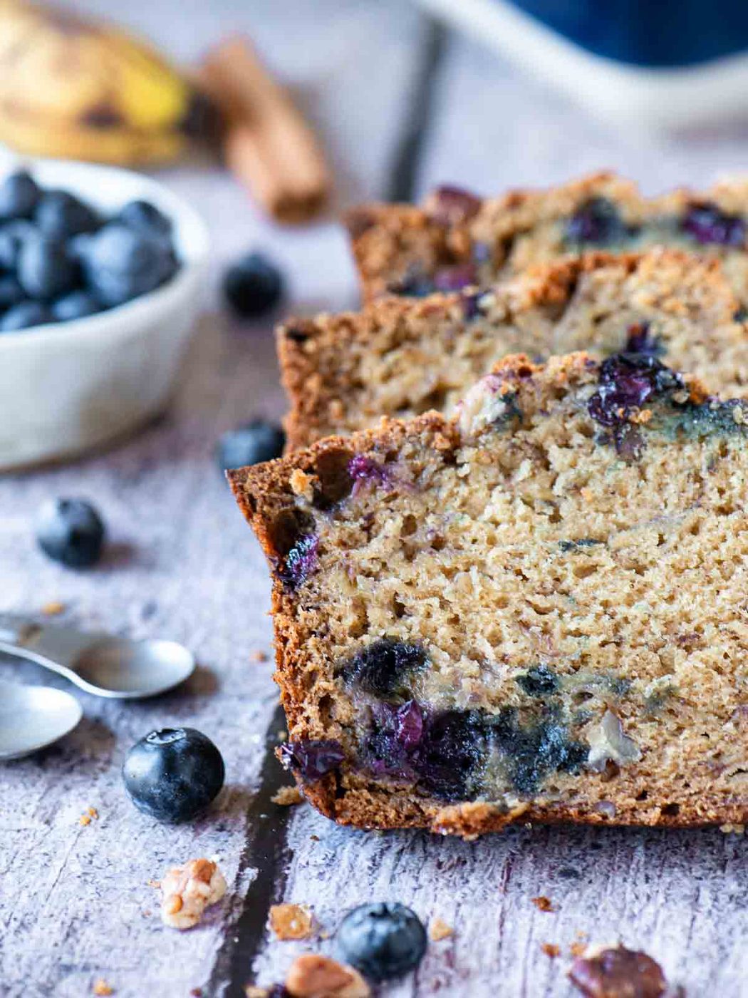 Three slices of blueberry banana bread on a wood table with fruit in the background.
