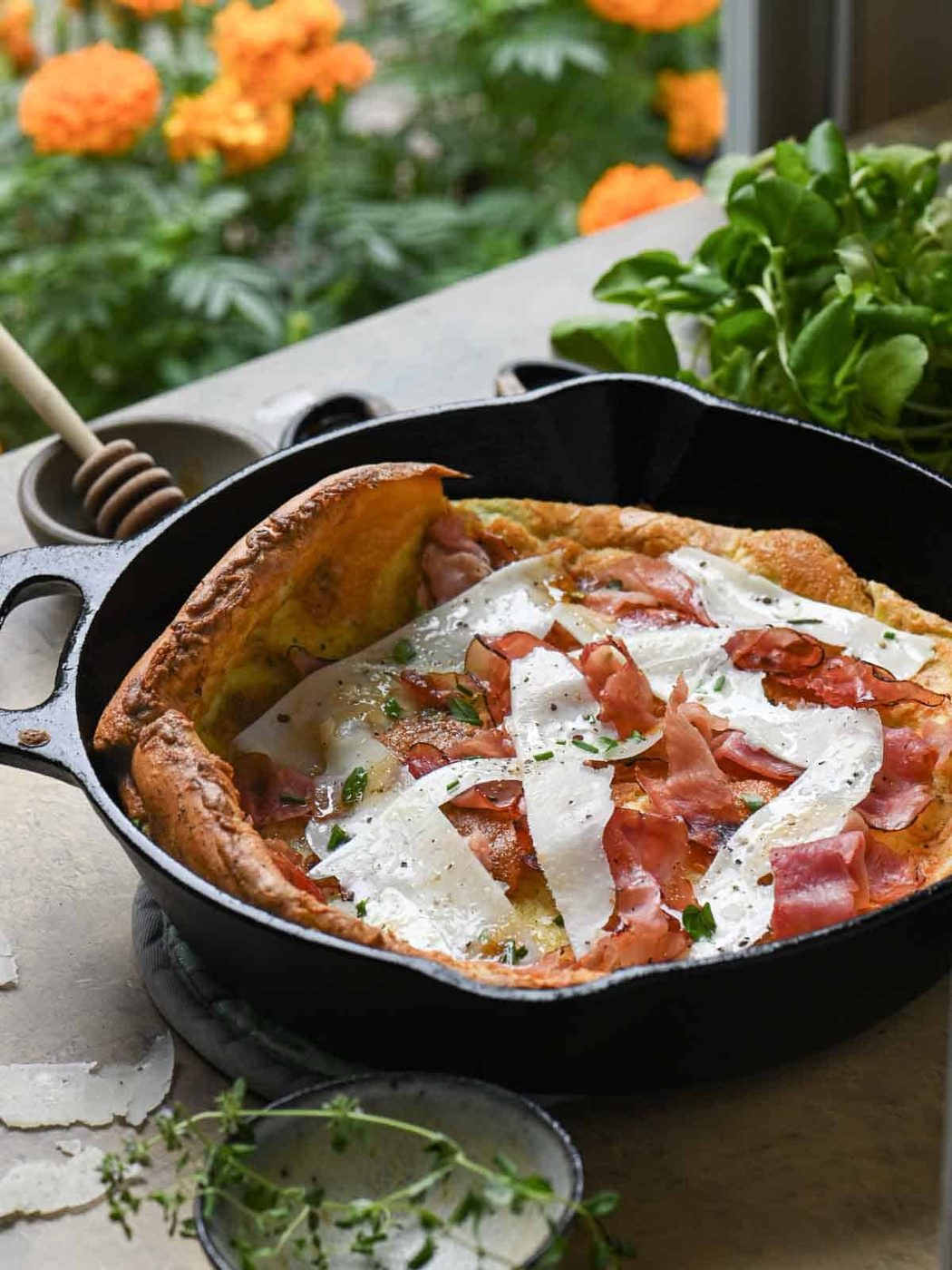 A Dutch baby pancake in a black skillet with greens and orange flowers in the background.