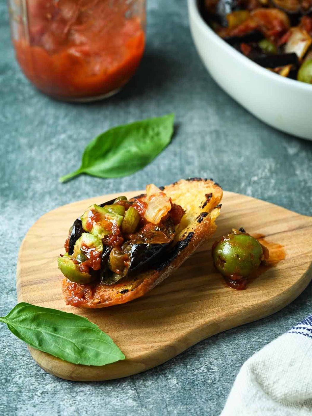 Piece of bread with caponata spread on it with basil and sauce on the side.