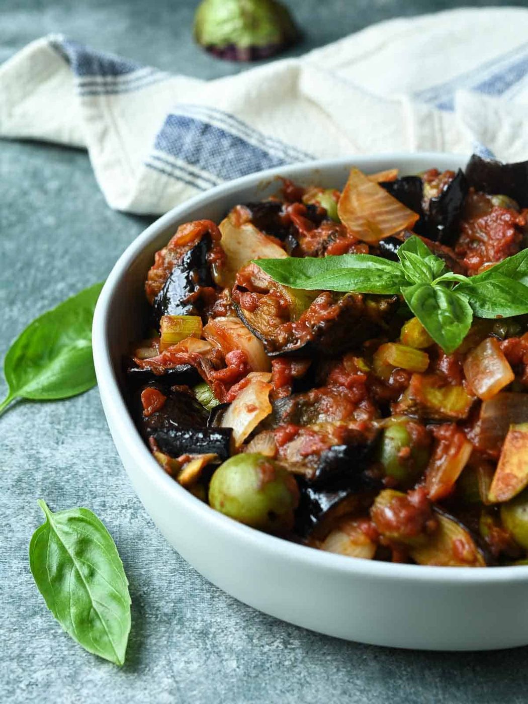 Eggplant caponata in a white bowl with basil leaves to the left.