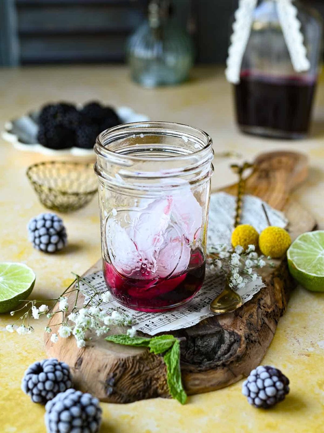 Ice and Blackberry syrup in a clear glass on top of a yellow surface with blackberries around it.