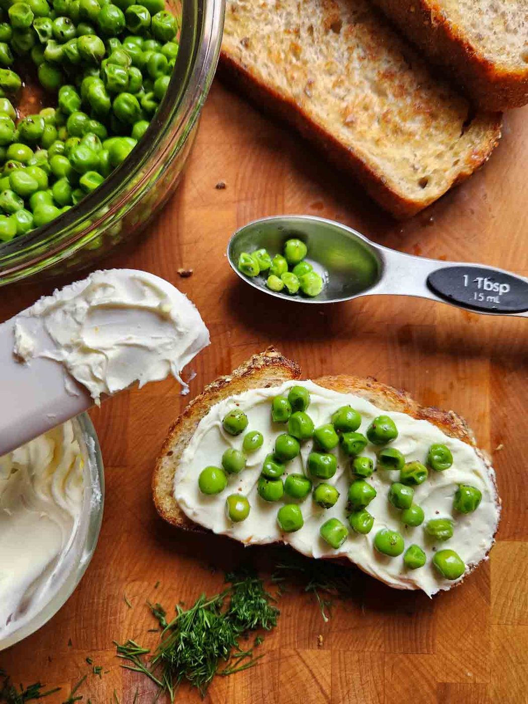 Assembling pea toast on a cutting board with mascarpone and dill to the side.