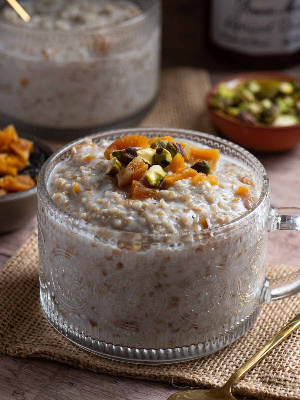 Close-up of apricot oatmeal in a clear mug with pistachios and apricots in the back.