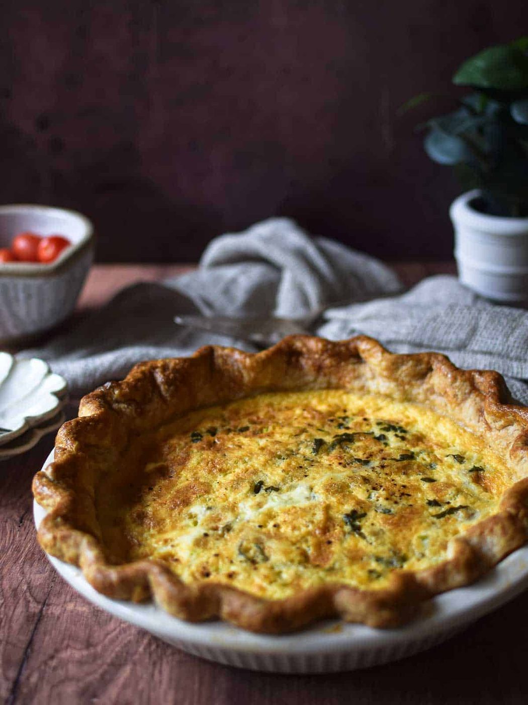 Angled view of tomato, leek and feta quiche on a brown surface with tomatoes and a linen in the background