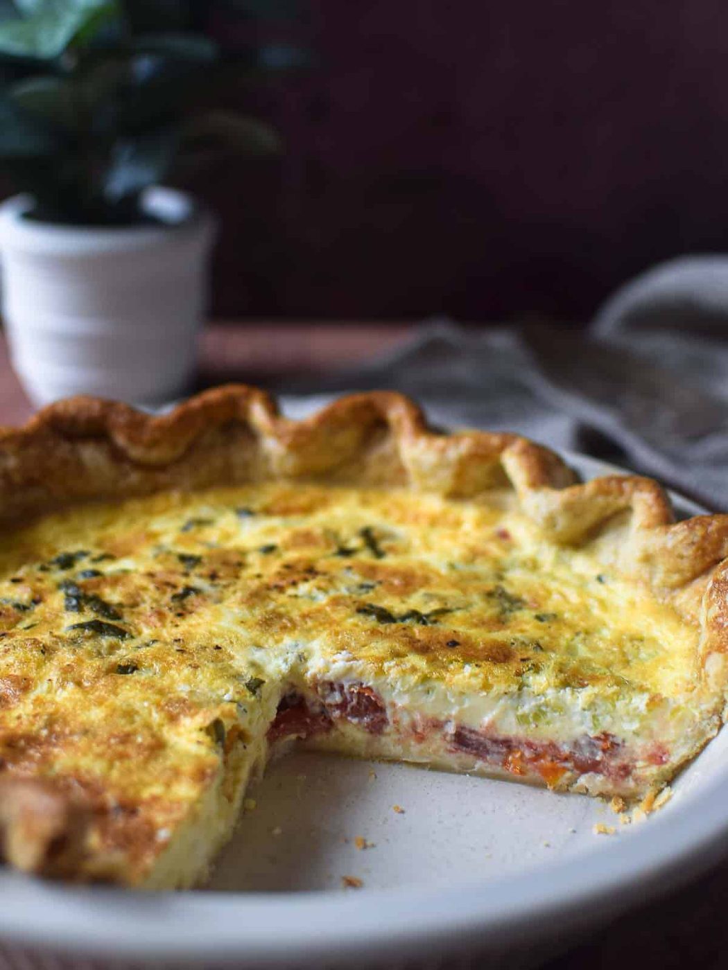 View of tomato, leek and feta quiche cut open so you can see the layers on a brown background and green plant in the background