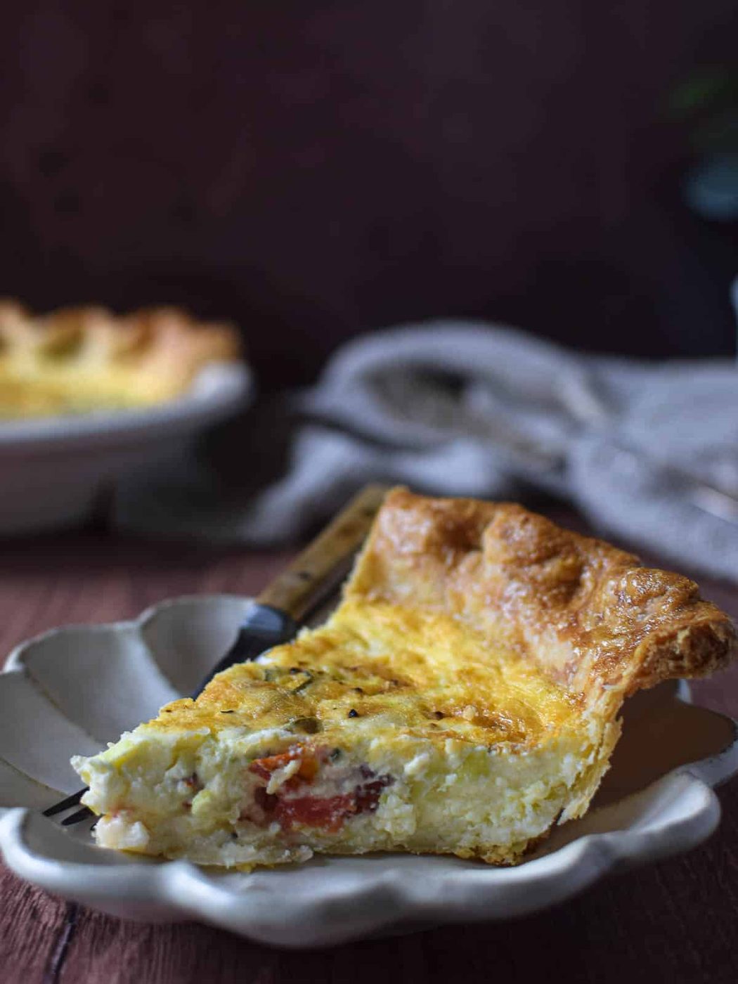 Slice of tomato, leek and feta quiche on a brown cutting board with quiche in the background