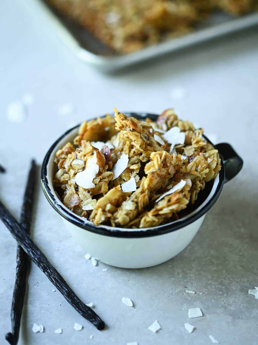 Vanilla coconut granola in a white antique cup with vanilla beans beside it.
