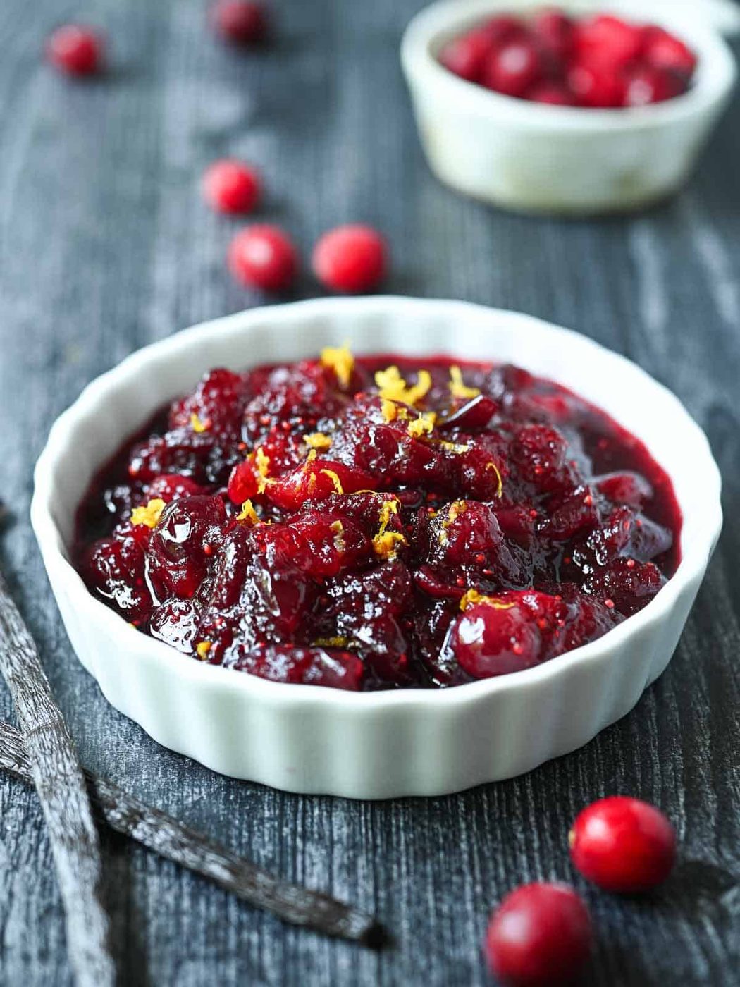 Cranberry compote in a white ramekin with cranberries and vanilla beans beside it.