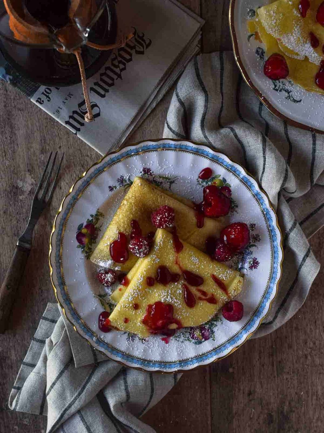 Overhead picture of stuffed crepes on a flowered plate with raspberry sauce on top on a wooden background.