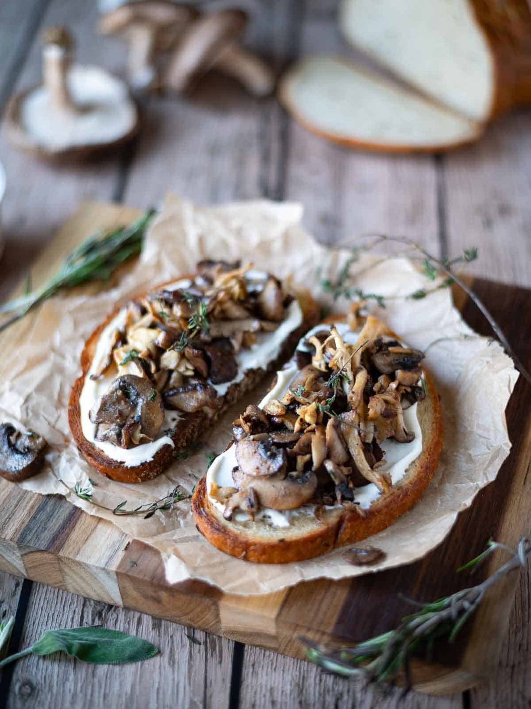Two slices of mushroom toast on a wood board with herbs around it.