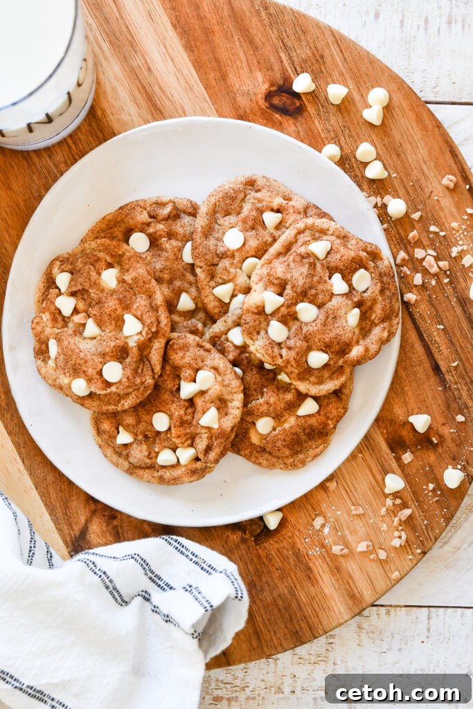 A close-up shot of several freshly baked, golden-brown snickerdoodle cookies, generously studded with white chocolate chips and toffee bits, resting on a cooling rack. The cookies are perfectly round, showcasing their inviting chewy texture and the sparkling cinnamon-sugar coating. One cookie is broken in half, revealing its soft, gooey interior with melted white chocolate.