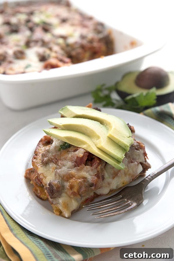 Beef enchilada casserole on a white plate in front of the pan. Slices of avocado on top. 