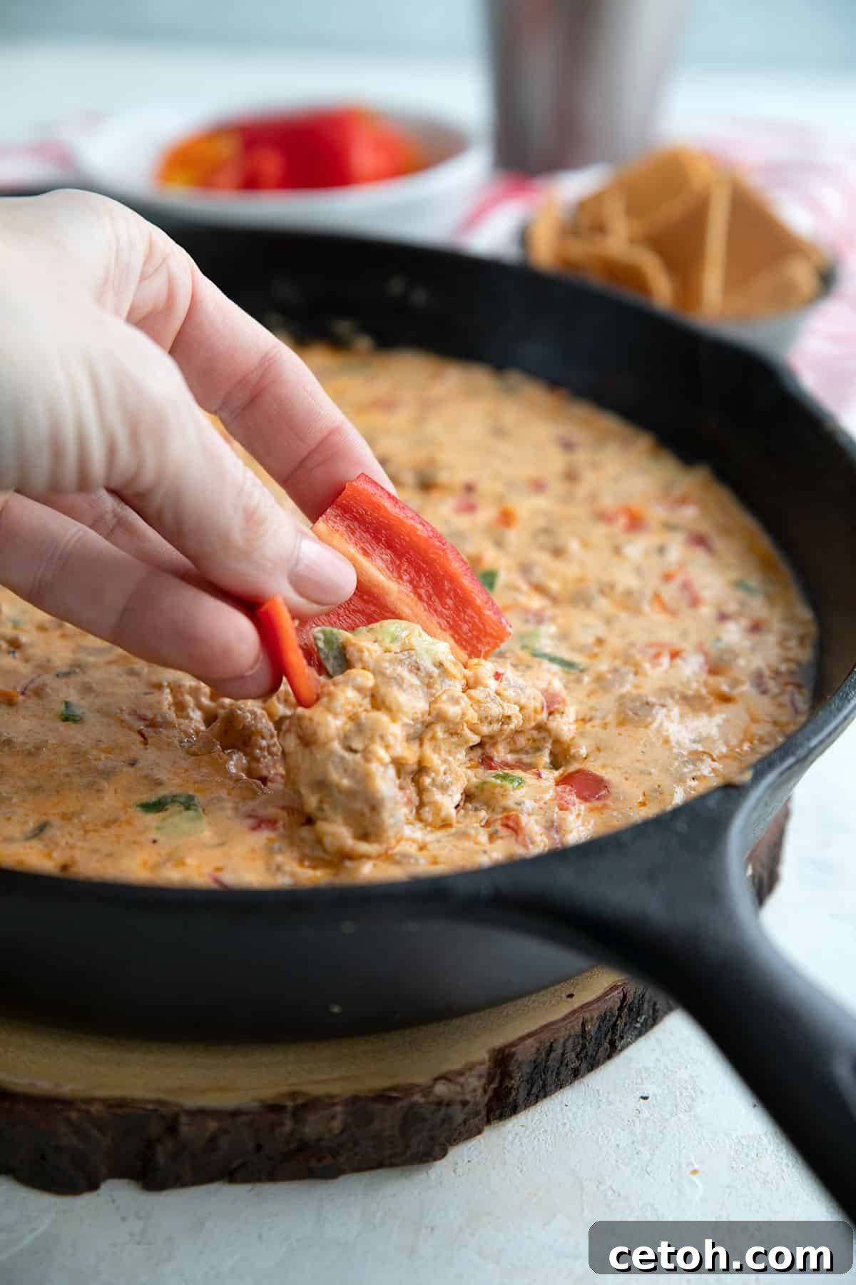 A hand dipping red pepper into cheesy sausage dip.