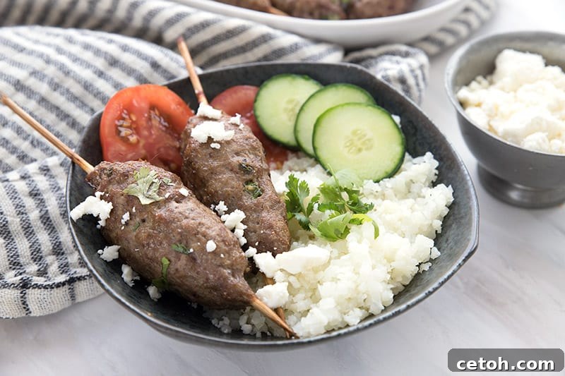 A black bowl showcasing beautifully cooked lamb kofta, served alongside fluffy cauliflower rice, vibrant cherry tomatoes, and crisp cucumber slices.