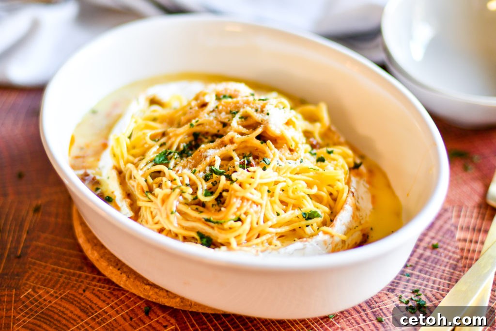 Creamy Baked Brie Spaghetti 4 Ingredients for Baked Brie Spaghetti laid out on a marble surface, including a wheel of brie, sun-dried tomatoes, roasted garlic cloves, fresh basil leaves, red pepper flakes, and uncooked spaghetti.