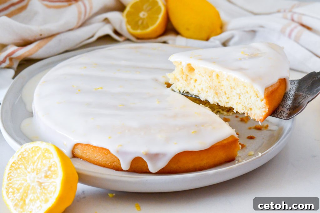 A close-up of a beautifully glazed lemon syrup cake, showing the smooth, slightly translucent white glaze with visible specks of lemon zest, ready for serving.