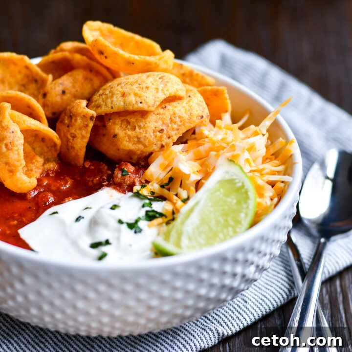 A vibrant bowl of Dad's Southern Homemade Chili, generously topped with shredded cheddar cheese, a dollop of sour cream, and a handful of crispy Fritos, is served on a rustic wooden surface, highlighting its hearty appeal.